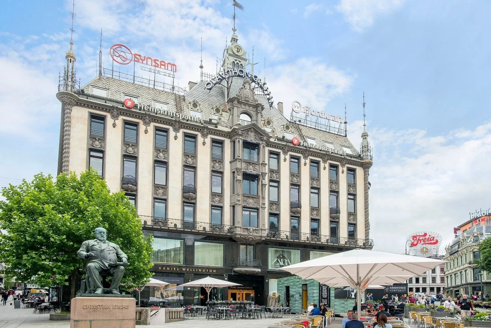Fasaden til Karl Johans gate 25 i Oslo, en lys bygård fra 1800-tallet med tårn og butikker i gateplan, sett fra Christian Krohgs statue på Egertorget.